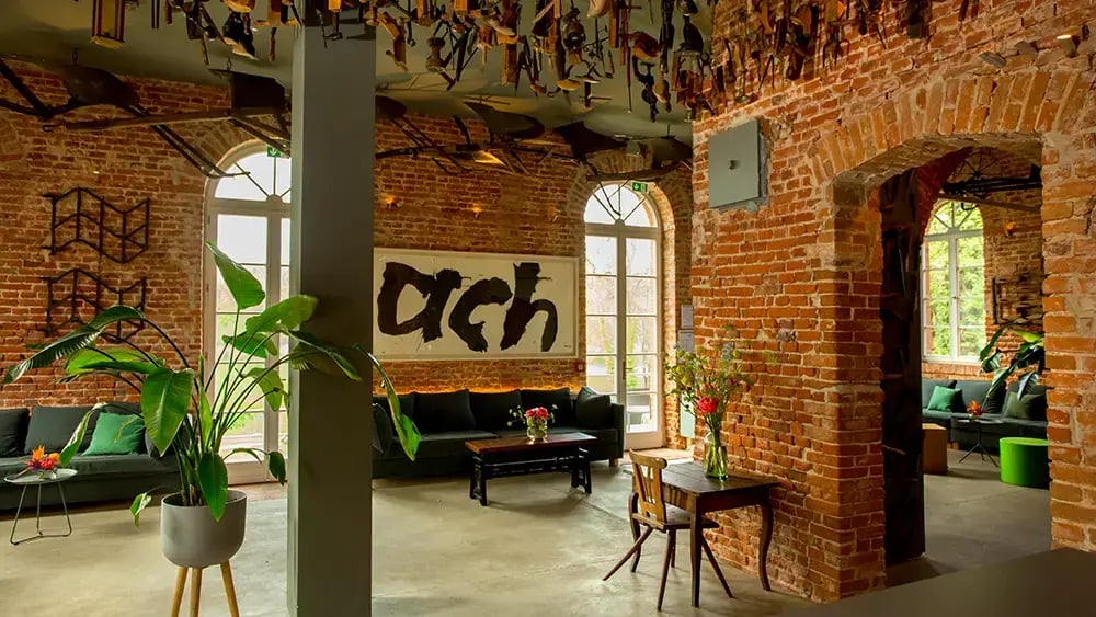 Offener Raum mit Backsteinwänden, großen Fenstern, Pflanzen, Tischen mit Stühlen und einem Sofa. An der Wand hängt ein Bild mit der Schrift 'ach'.