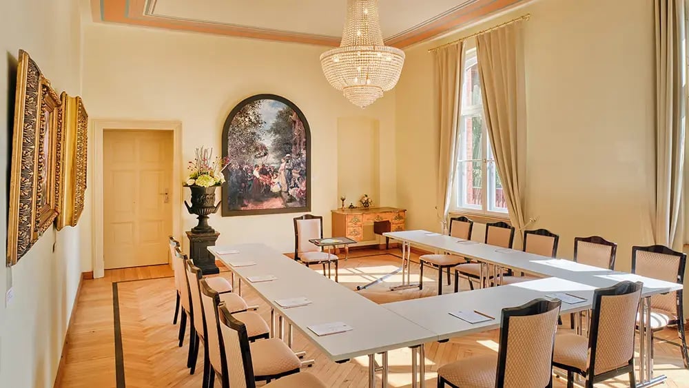 Heller Konferenzraum mit U-förmigem Tisch, gepolsterten Stühlen, Kronleuchter, großen Fenstern und klassischer Wanddekoration.
