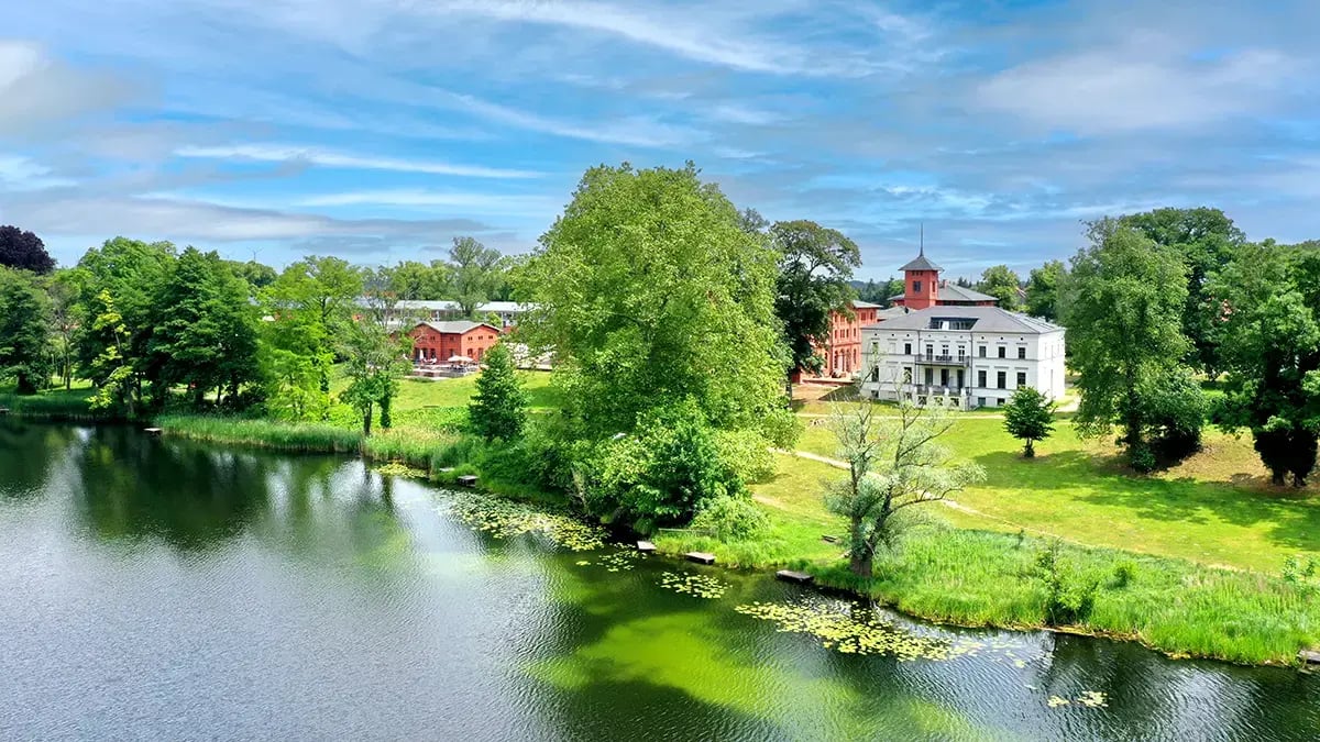 Schloss mit weißem Hauptgebäude und rotem Turm neben See mit grünen Seerosen und Bäumen unter blauem Himmel mit Wolken.