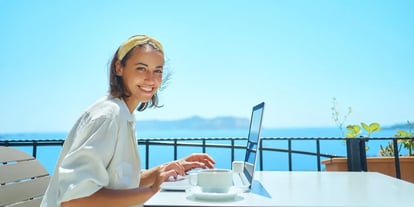 Lächelnde Frau sitzt mit Laptop an einem Tisch auf der Terrasse eines Hotels oder Restaurants, im Hintergrund blauer Himmel und Meer – Symbolbild für Blended Travel, bei dem Geschäftsreisende Arbeit und Freizeit miteinander verbinden, etwa im Rahmen von Bleisure- oder Workation-Aufenthalten nach einem MICE-Event oder einer Geschäftsreise.