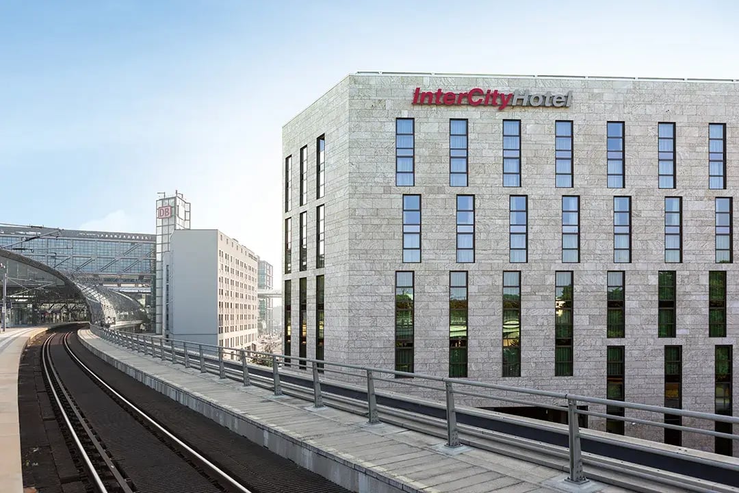 Ein modernes, helles Gebäude mit rechteckigen Fenstern steht direkt an einer erhöhten Bahntrasse. An der Fassade ist das Logo 'InterCityHotel' in roten und grauen Buchstaben angebracht. Im Hintergrund sind Glas- und Stahlkonstruktionen des Berliner Hauptbahnhofs zu sehen. Der Himmel ist klar.