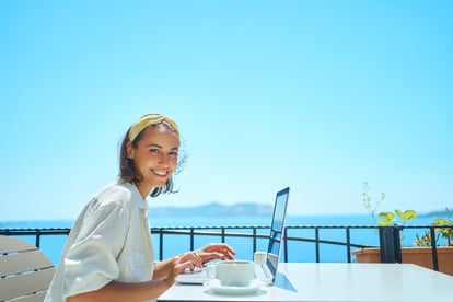 Lächelnde Frau sitzt mit Laptop an einem Tisch auf der Terrasse eines Hotels oder Restaurants, im Hintergrund blauer Himmel und Meer – Symbolbild für Blended Travel, bei dem Geschäftsreisende Arbeit und Freizeit miteinander verbinden, etwa im Rahmen von Bleisure- oder Workation-Aufenthalten nach einem MICE-Event oder einer Geschäftsreise.