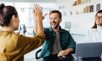 Two colleagues in business attire high-five each other with beaming smiles in a modern, bright office; in the background, additional desks and workstations are visible—a symbolic image of the moment of triumph following successful budget negotiations in event management. The gesture symbolizes efficient teamwork, mutual appreciation, and the positive results that strategic MICE management enables despite budget pressures and rising costs: consolidated requests, negotiated terms, and greater impact with the same or a smaller budget.