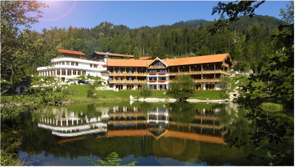 Mehrstöckiges Hotel im alpenländischen Landhausstil mit Holzbalkonen und ziegelrotem Dach, das sich in einem ruhigen See spiegelt. Im Hintergrund erstreckt sich dichter Mischwald an einem Berghang, im Vordergrund Schilfgras und Bäume bei sonnigem Wetter mit blauem Himmel.