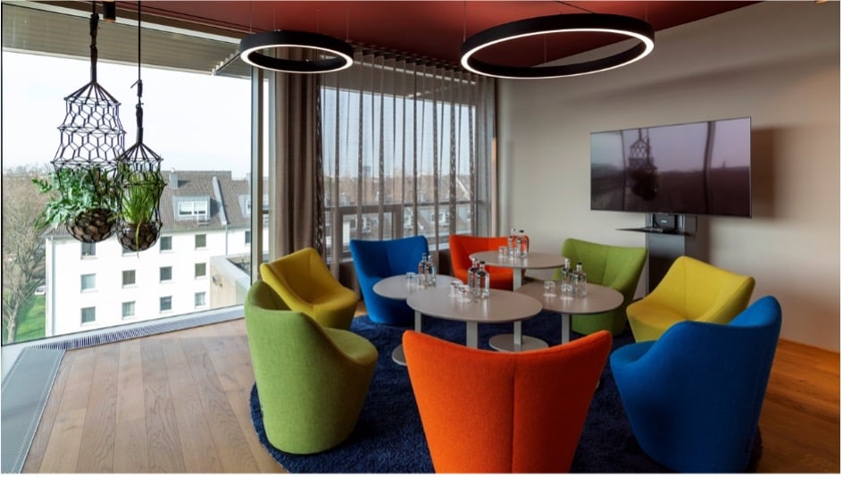 Moderner Lounge-Meetingbereich mit bunten Designsesseln in Grün, Gelb, Orange und Blau, gruppiert um zwei runde Tische auf einem dunkelblauen Teppich. Im Hintergrund ein wandmontierter Großbildschirm und große Fenster mit Ausblick über die Stadt. An der Decke runde LED-Ringleuchten und hängende Pflanzen in Makramee-Ampeln auf einem Holzboden.