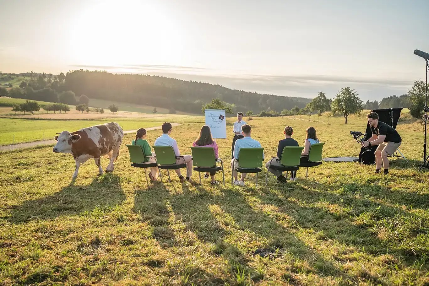 Outdoor meeting at DEKRA Congresshotel Wart in the Black Forest. A small group of seminar participants sits on a grassy meadow while a presenter leads the session. A photographer captures the moment from the side, and a cow walks past nearby. In the background, rolling fields and the forests of the Black Forest are bathed in the warm light of the evening sun.