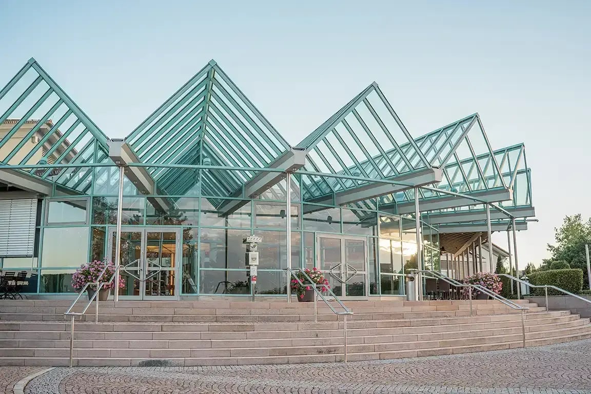 Modern exterior view of the DEKRA Congresshotel Wart conference and event hotel in Germany’s Black Forest. The glass façade reflects the surrounding landscape, while a staircase leads to multiple entrances featuring contemporary metal design elements. Planted flower containers are placed beside the entrances.
