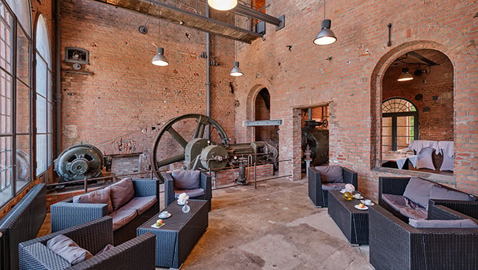 Innenansicht eines historischen Veranstaltungsraums im Landgut Stober: offener Raum mit hohen Backsteinwänden, Rundbogenarkaden und industriellen Elementen wie einer original erhaltenen Maschine („Dicke Bertha“). Moderne Lounge-Möbel stehen im Kontrast zur historischen Architektur im Loft- und Gewölbestil. Große Fensterflächen lassen viel Tageslicht einfallen und unterstreichen den besonderen Charakter der nachhaltigen Tagungslocation.