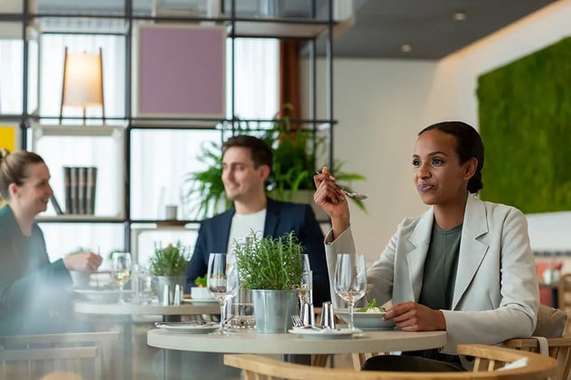 Helle Restaurant-Szene mit modernem Interieur, runden Holztischen und grünen Pflanzen als Tischdekoration. Im Hintergrund sind Regale mit quadratischen, farbigen Flächen und eine große, grüne Wandverkleidung zu sehen. Auf den Tischen stehen Weingläser, Wassergläser und weißes Geschirr. Eine Person im Vordergrund trägt ein helles Sakko und hält eine Gabel in der rechten Hand.