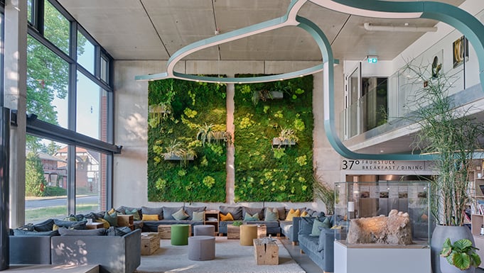 Moderne Lobby und Aufenthaltsbereich im Landgut Stober mit großer Fensterfront und Blick ins Grüne: heller Raum mit minimalistischer Architektur, kombiniert mit einer großflächigen vertikalen Pflanzenwand (Mooswand) als zentrales Designelement. Sitzlandschaften aus Sofas und Hockern schaffen eine entspannte Atmosphäre für Gäste und Veranstaltungsteilnehmende. Die Verbindung aus nachhaltigem Design, natürlichem Grün und modernem Innenausbau unterstreicht den Charakter der Location als innovative Tagungs- und Eventlocation mit Fokus auf Nachhaltigkeit und Aufenthaltsqualität.