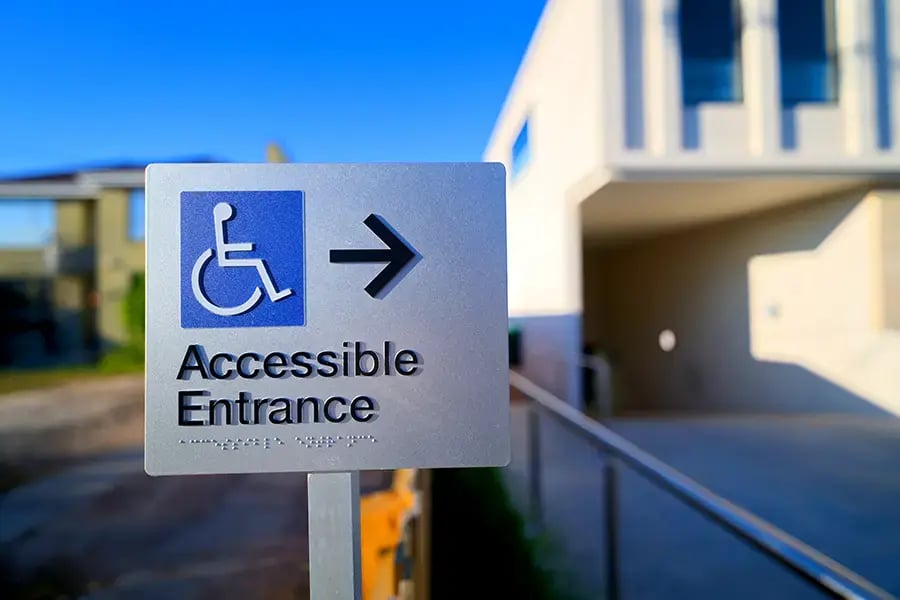 Schild mit Rollstuhlsymbol und Pfeil nach rechts, darunter der Text 'Accessible Entrance' und die Beschriftung mit Blindenschrift.