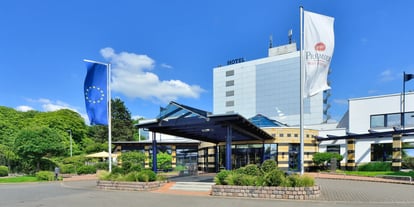 Im Vordergrund steht der Eingangsbereich eines modernen Hotelgebäudes mit einer Glasüberdachung. Links und rechts sind zwei Fahnenmasten zu sehen, einer mit einer Europaflagge und einer mit einer weißen Fahne, auf der das Logo von Best Western Premier zu erkennen ist. Das Hauptgebäude im Hintergrund ist mehrstöckig, hat eine silbergraue Fassade und trägt oben das Wort 'HOTEL'. Rechts neben dem Haupteingang befindet sich ein niedrigerer Gebäudeteil mit gelb-weißen Streifen. Die Umgebung ist gepflegt, mit gepflasterten Wegen, Rasenflächen und Bäumen.