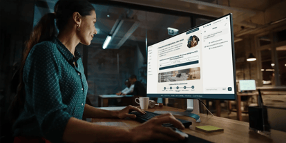 Person sitzt an einem Schreibtisch und arbeitet mit der neuen Oberfläche des MICE Portals auf einem großen Monitor in einem modernen Büro mit weiteren Arbeitsplätzen im Hintergrund.