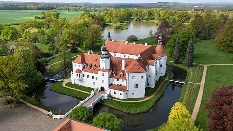 Luftaufnahme eines weißen Schlosses mit roten Dächern, umgeben von Wassergräben und grünen Bäumen.