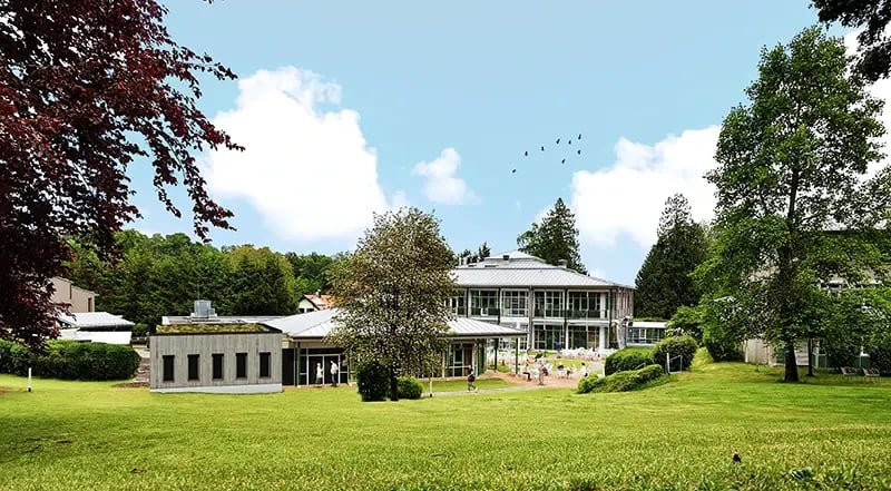 Seminarhaus mit mehreren Gebäudeteilen, umgeben von Rasenfläche und Bäumen unter blauem Himmel mit Wolken und fliegenden Vögeln.