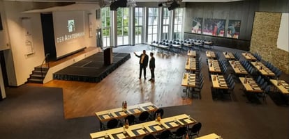 Großer Veranstaltungssaal mit Holzboden, Bühne mit Leinwand 'Der Achtermann', zwei Personen sprechen in der Mitte, Reihen von Tischen und Stühlen