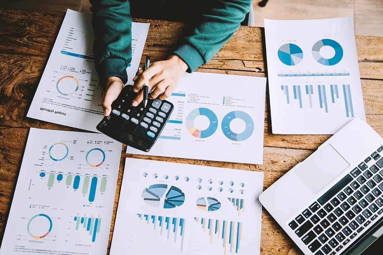 Person mit grünem Pullover bedient Taschenrechner auf Holztisch mit mehreren Blättern voller bunter Kreis- und Balkendiagramme neben aufgeklapptem Laptop.