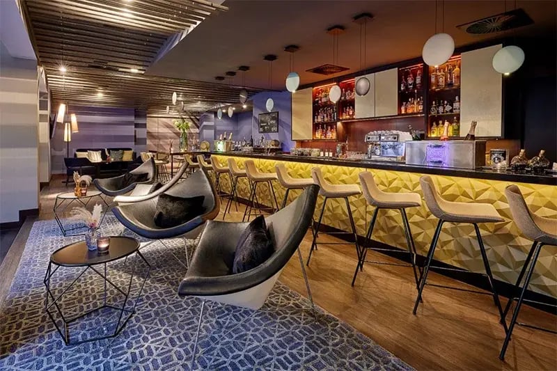 Moderne Bar mit gelben Barhockern, schwarzen Sesseln, blauem Teppich und beleuchtetem Tresen mit Spirituosenregalen im Hintergrund.