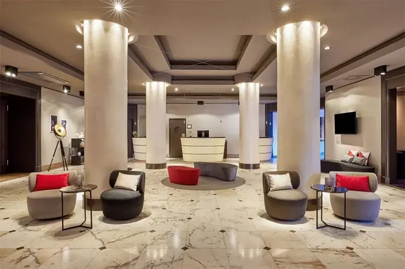 Helle Hotellobby mit Marmorboden, runden Säulen, Sitzgruppen mit grauen und roten Sesseln und einem Empfangstresen im Hintergrund.