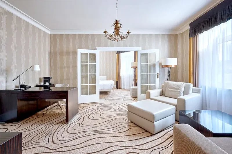 Helles Wohnzimmer mit Sessel, Hocker, Schreibtisch und offenem Durchgang zu Schlafzimmer mit Doppelbett, große Fenster mit Vorhängen.