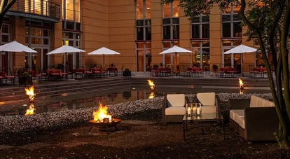 Außenbereich eines Hotels in Dresden mit Sitzgruppe, brennenden Feuerstellen und Tischen unter weißen Sonnenschirmen am Abend
