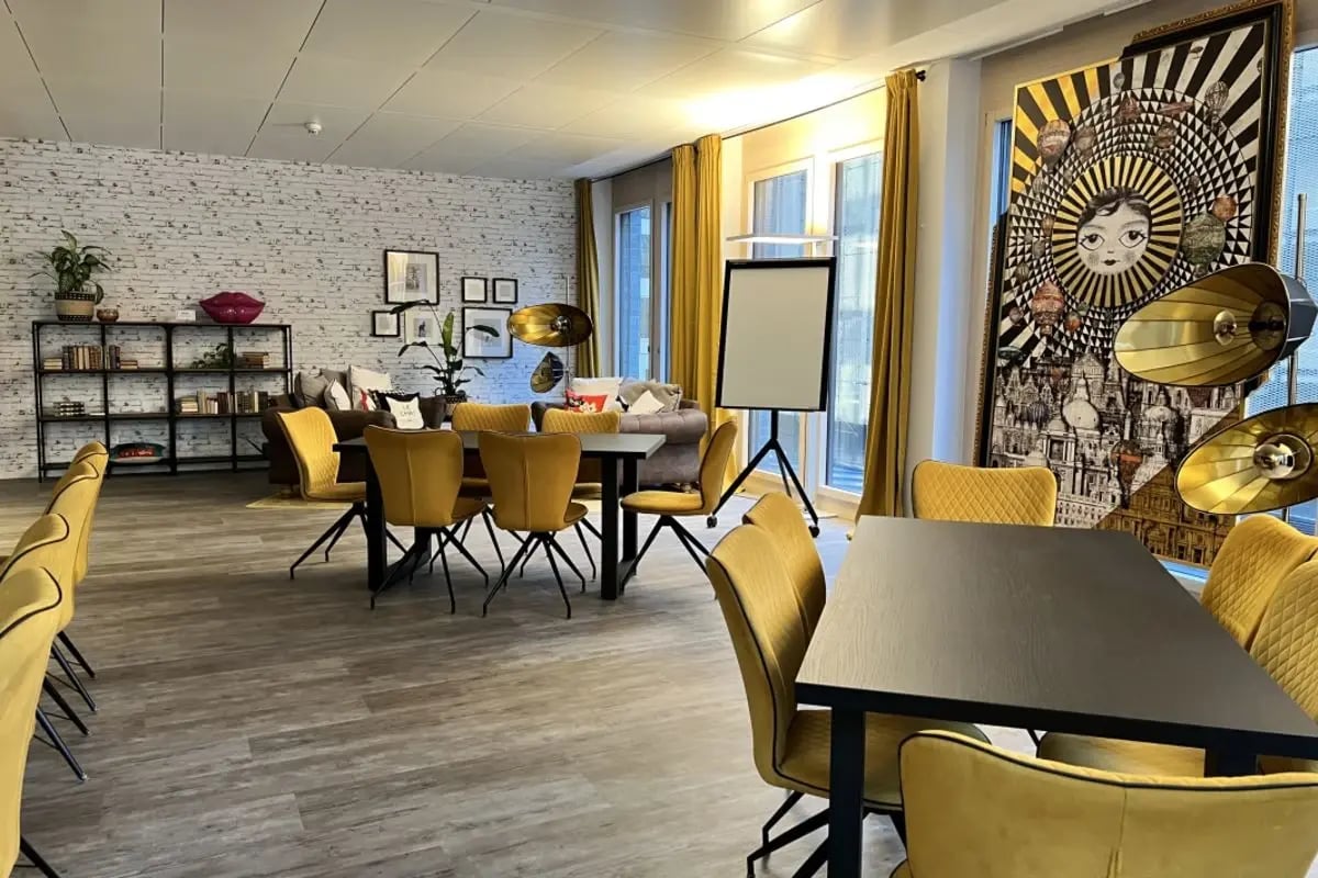 Moderner Besprechungsraum mit gelben Stühlen, schwarzen Tischen, großem Wandbild mit Sonnenstrahlenmuster und Flipchart.