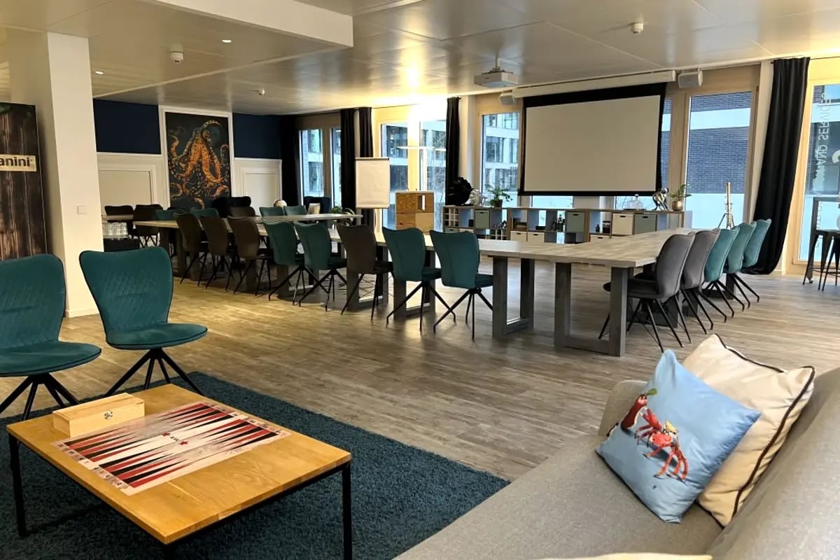 Großer Konferenzraum mit langem Holztisch, mehreren Stühlen, Projektionsleinwand und gemütlicher Sitzecke mit Sofa und Backgammon-Spiel auf dem Tisch.