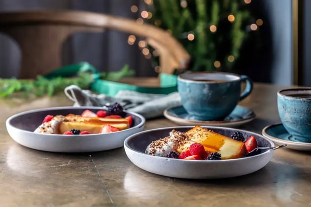 Zwei Teller mit Karamellpudding, garniert mit Beeren und Sahne, auf einem Tisch mit zwei blauen Tassen im Hintergrund.