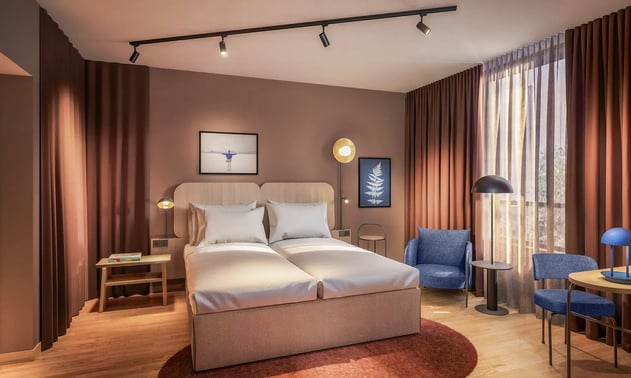 Hotelzimmer mit Doppelbett, zwei blauen Sesseln, braunen Vorhängen und zwei Bildern an der Wand.