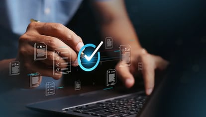 Hand tippt auf Laptop-Tastatur, darüber schwebt ein blaues Häkchen-Symbol mit mehreren transparenten Dokumenten-Icons