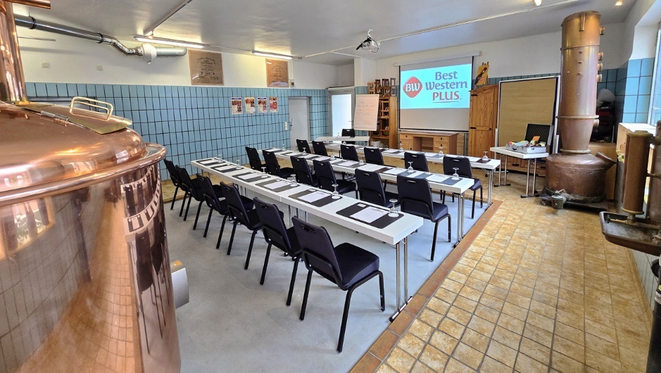Außergewöhnlicher Tagungsraum in einer historischen Brauerei mit parlamentarischer Bestuhlung in U-Form, dunklen Stühlen und weißen Tischen. Im Vordergrund ein großer kupferner Braukessel, im Hintergrund eine Leinwand mit Best Western Plus Logo, ein Flipchart und ein weiterer Kupferapparat. Die Wände sind mit hellblauen Fliesen verkleidet, der Boden besteht aus gelben Fliesen.