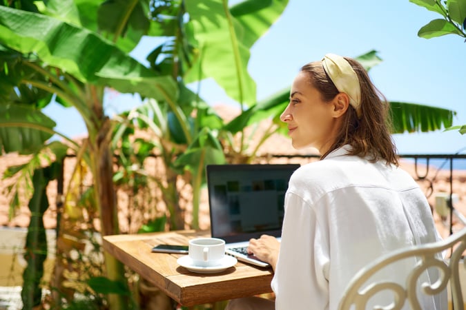 Dieselbe Frau wie im Header-Bild sitzt erneut am Laptop, daneben Smartphone und eine Kaffeetasse, auf dem Bildschirm ist im Hintergrund ein Kalender, vermutlich Outlook, zu erkennen, sie blickt jedoch lächelnd zur Seite statt auf den Bildschirm, im Hintergrund Bananenpalmen, rote Dachziegel und ein kleiner Meeresausblick, Symbolbild für Blended Travel und die entspannte Seite des mobilen Arbeitens zwischen Terminen und Freizeit.