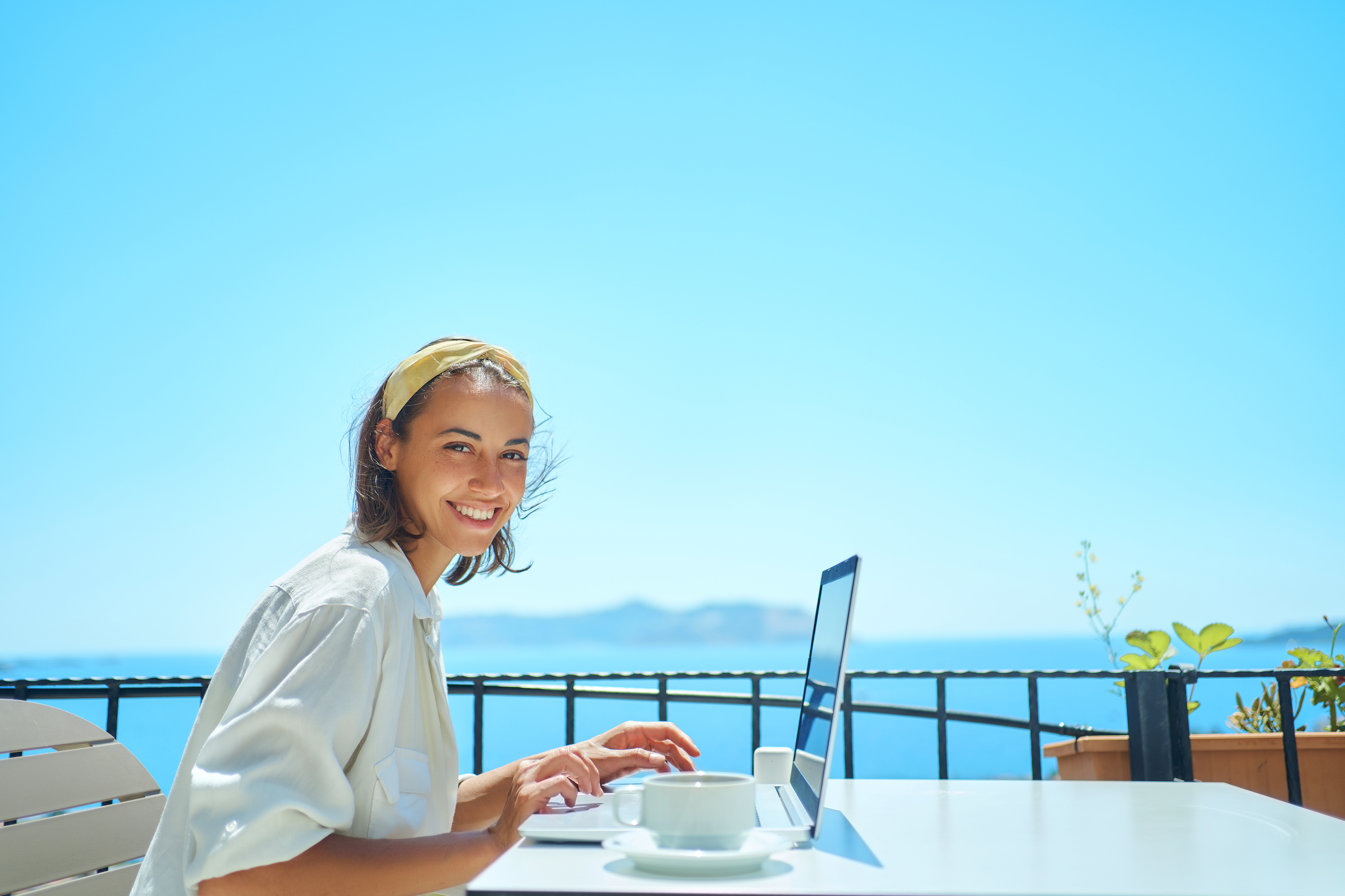 Lächelnde Frau sitzt mit Laptop an einem Tisch auf der Terrasse eines Hotels oder Restaurants, im Hintergrund blauer Himmel und Meer – Symbolbild für Blended Travel, bei dem Geschäftsreisende Arbeit und Freizeit miteinander verbinden, etwa im Rahmen von Bleisure- oder Workation-Aufenthalten nach einem MICE-Event oder einer Geschäftsreise.