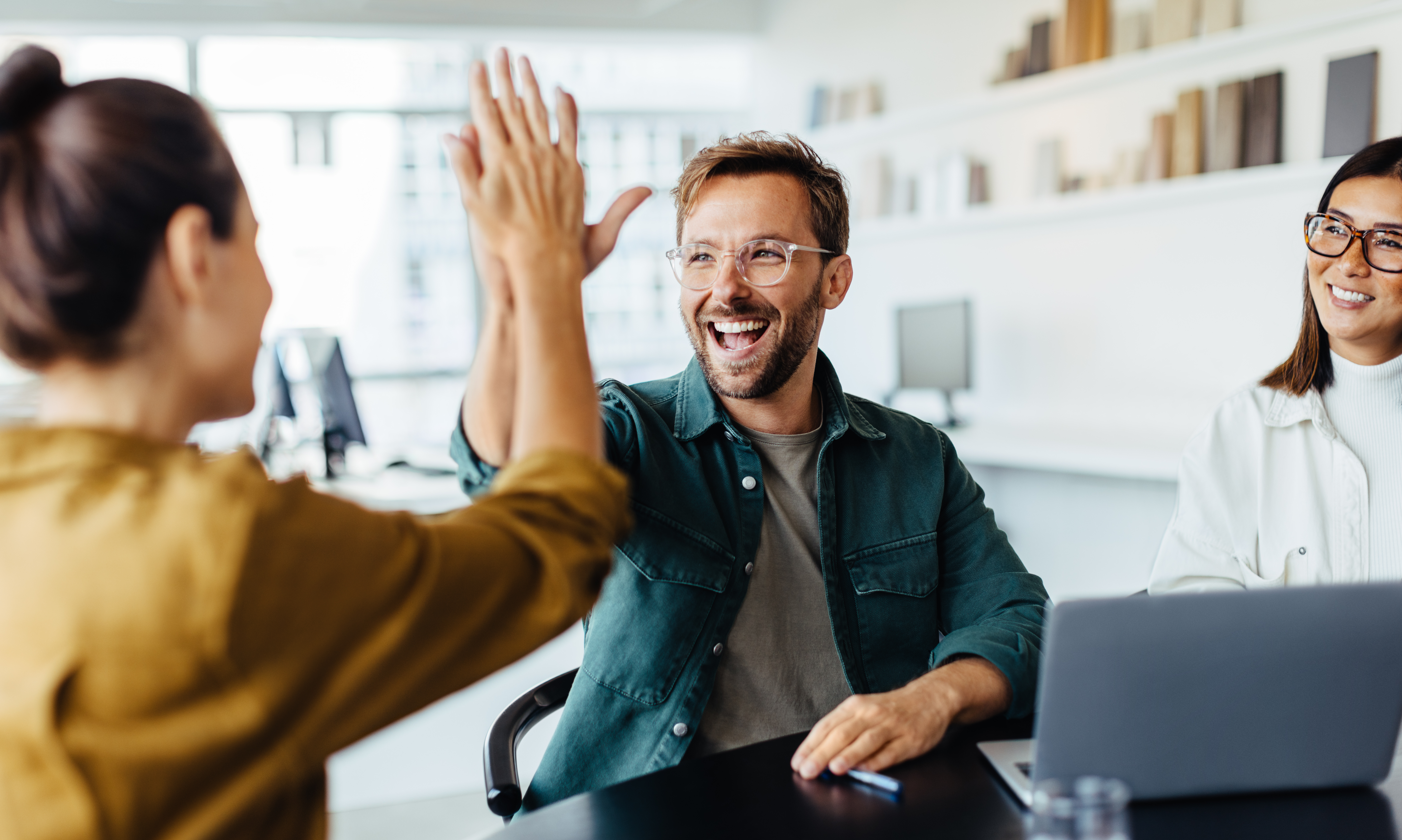 Zwei Kollegen in Business-Kleidung klatschen sich freudestrahlend in einem modernen, hellen Büro ab, im Hintergrund sind weitere Schreibtische und Arbeitsplätze zu erkennen – ein Symbolbild für den Erfolgsmoment nach gelungener Budgetverhandlung im Eventmanagement. Die Geste steht sinnbildlich für effiziente Teamarbeit, gegenseitige Wertschätzung und die positiven Ergebnisse, die strategisches MICE Management trotz Budgetdruck und steigender Kosten ermöglicht: gebündelte Anfragen, verhandelte Konditionen und mehr Wirkung bei gleichem oder kleinerem Budget