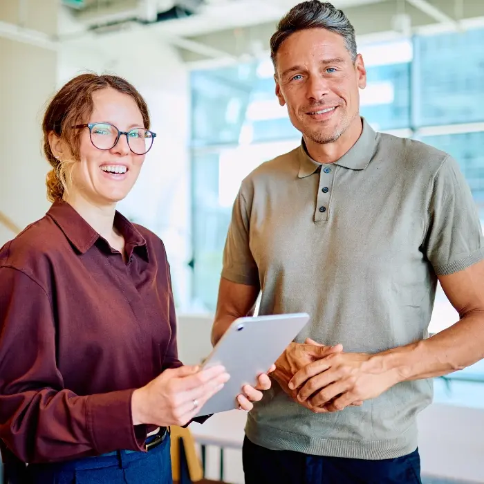Mann und Frau stehen lächelnd nebeneinander im Büro, die Frau hält ein Tablet, symbolisch für die Phase Verankern im Strategischen MICE Management.