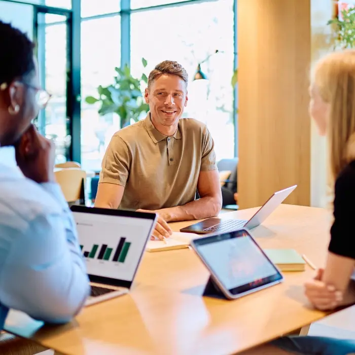 Team im Gespräch am Tisch mit Laptops und Kennzahlen, ein lächelnder Mann im Austausch mit Kolleginnen, symbolisch für die Phase Weiterentwickeln im Strategischen MICE Management.