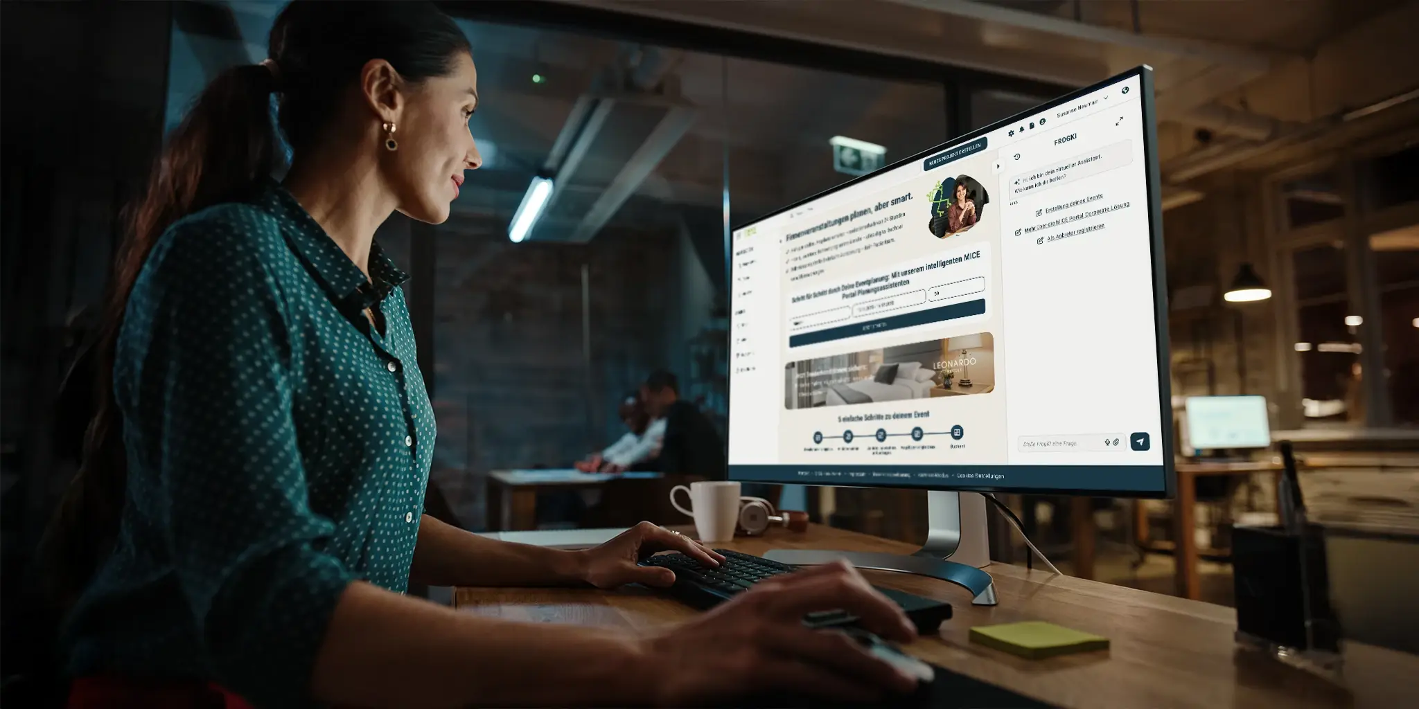 Person sitzt an einem Schreibtisch und arbeitet mit der neuen Oberfläche des MICE Portals auf einem großen Monitor in einem modernen Büro mit weiteren Arbeitsplätzen im Hintergrund.