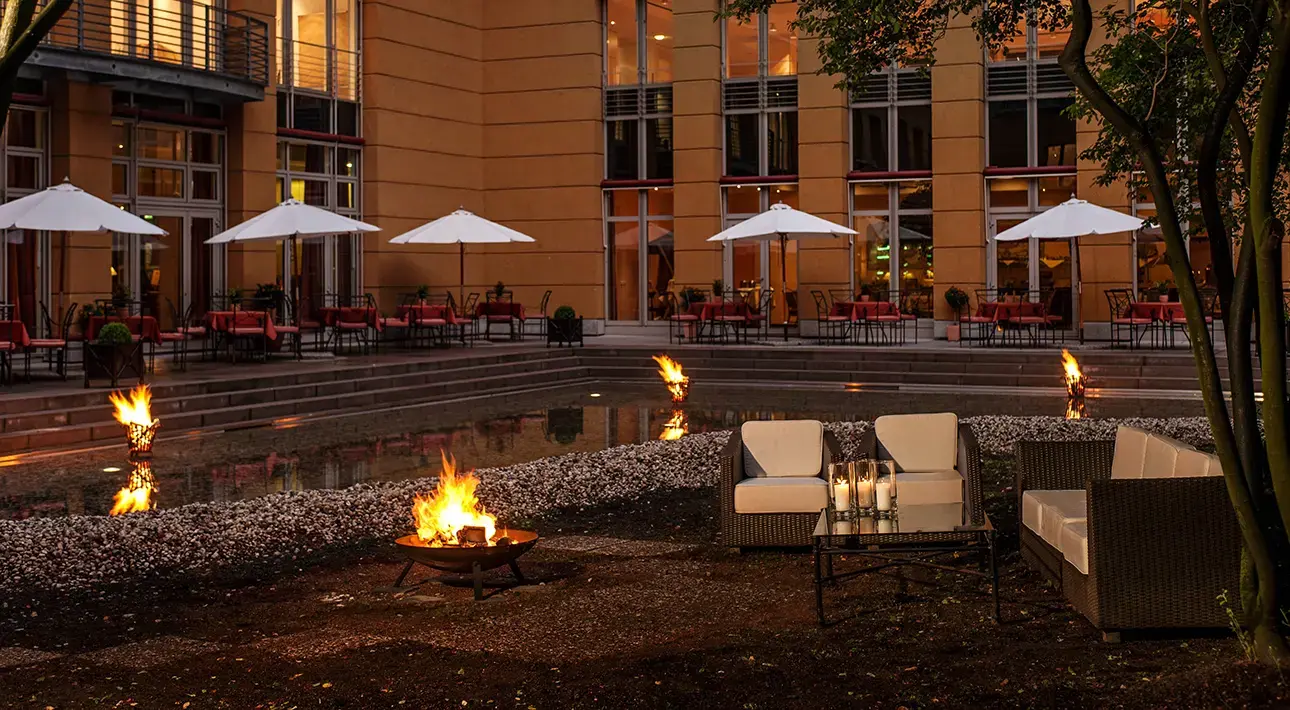 Außenbereich eines Hotels in Dresden mit Sitzgruppe, brennenden Feuerstellen und Tischen unter weißen Sonnenschirmen am Abend