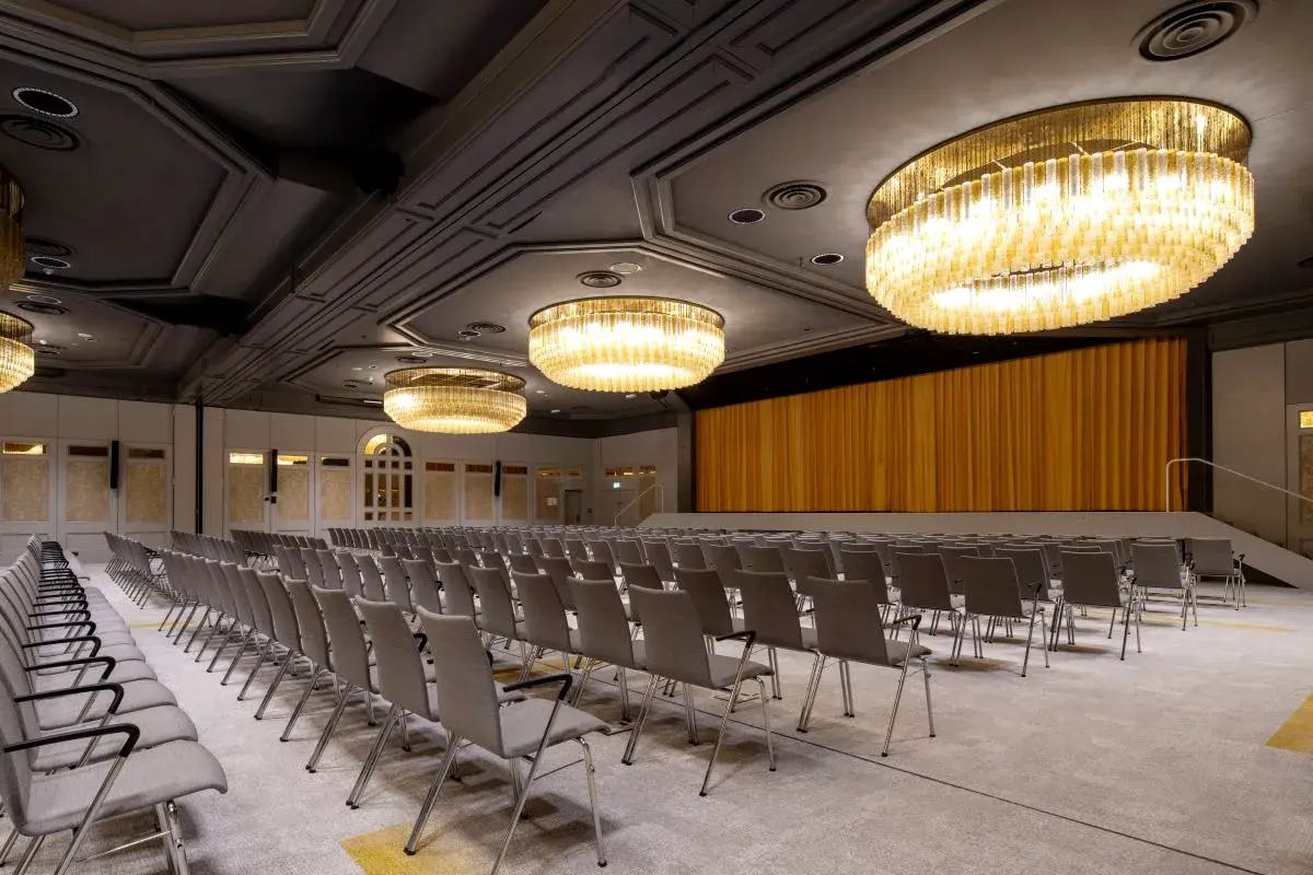 Ballsaal mit mehreren Reihen grauer Stühle, zwei großen Kristalllüstern an der Decke und einem goldenen Vorhang auf der Bühne.